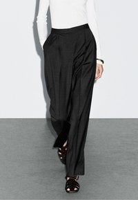 Zwarte geplooide broek van een textiel met structuur en een hoge tailleband, gecombineerd met donkere sandalen met bandjes, gedragen over een getailleerd wit topje.