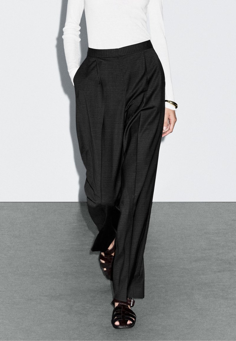 Zwarte geplooide broek van een textiel met structuur en een hoge tailleband, gecombineerd met donkere sandalen met bandjes, gedragen over een getailleerd wit topje.