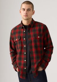 Rode en groene geblokte flanellen shirt met knoopsluiting, twee borstzakken en een relaxte pasvorm, gedragen over een zwart T-shirt.