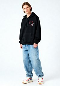 Felpa nera con cappuccio e tasca frontale, decorata con un ricamo a forma di cuore rosso, abbinata a jeans baggy blu chiaro e sneakers bianche con motivo.