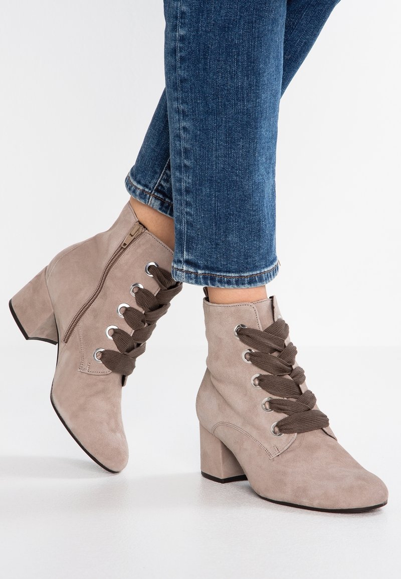 Botines de ante beige con tacón apilado, que presentan un cierre lateral y cordones marrones que se entrelazan a través de ojales plateados.