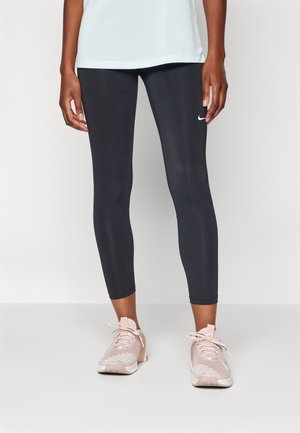 Donna che indossa leggings neri Nike e scarpe sportive rosa chiaro, in piedi contro uno sfondo bianco uniforme.