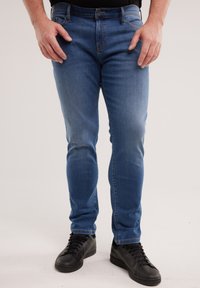 Mees, kes kannab kitsaid siniseid teksapükse ja musti tenniseid, seisab käed lõdvalt külgedel ühetaustal.