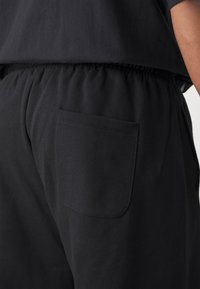 Schwarze Jogginghosen aus weichem Stoff verfügen über einen elastischen Bund und eine einzelne Gesäßtasche. Die Struktur wirkt glatt, ohne sichtbare Muster.