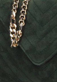 Donkergroene suède handtas met een gequilt chevronpatroon, versierd met een glanzende gouden schouderband voor extra detail.