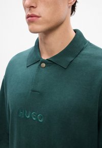 Tumed roheline polo särk pehmest puuvillast, millel on soonikkoes krae, nupukinnitus ja rinnal broderitud "HUGO" logo.