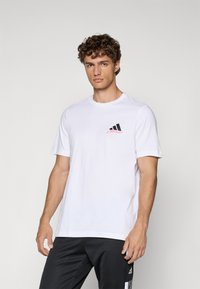Hvid bomulds t-shirt med korte ærmer, rund halsudskæring, med et sort og lyserødt logo på venstre bryst. Sammen med mørkegrå bukser.