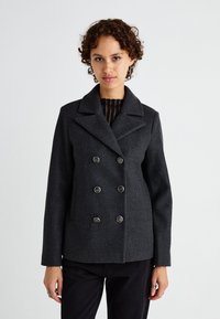 Cappotto in lana grigio scuro con design doppiobotte, grandi bottoni, colletto a bavero e tasche laterali. Modello che indossa un top a righe sotto.