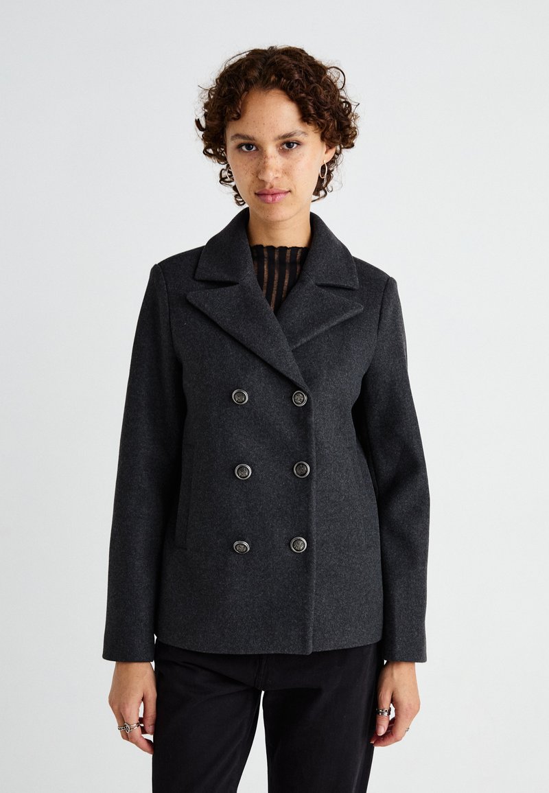 Cappotto in lana grigio scuro con design doppiobotte, grandi bottoni, colletto a bavero e tasche laterali. Modello che indossa un top a righe sotto.