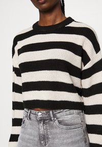 Černý a krémový pruhovaný pletený svetr s krátkým střihem a kulatým výstřihem, doplněný vybledlými šedými džínami s knoflíkovým zapínáním.
