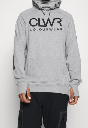Person trägt einen grauen Kapuzenpullover mit dem Aufdruck "CLWR COLOURWEAR" und schwarze Cargohosen, steht vor einem schlichten weißen Hintergrund.