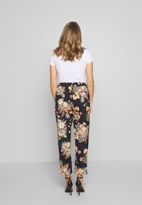 Vrouw die met de rug naar de camera staat, in een wit T-shirt, zwarte bloemenbroek en zwarte hoge hakken, tegen een effen grijze achtergrond.