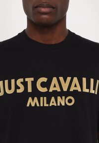 Černé tričko s zlatým textem "JUST CAVALLI MILANO." Má kulatý výstřih a krátké rukávy, je vyrobeno z měkké látky s hladkým povrchem.