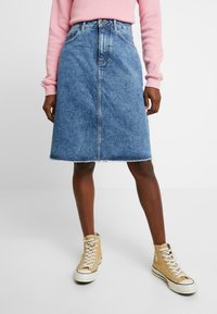 Denim A-linje kjol i medelblått med en bleknad effekt, med knappstängning fram och fransad nederkant. Matchas med beige högstövlar.