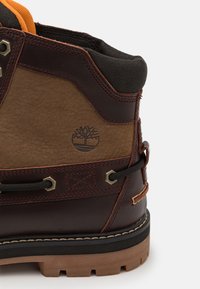 Brown leather boot with dark brown accents and orange lining. Features a textured surface, metal eyelets, and a rubber sole with distinct tread.