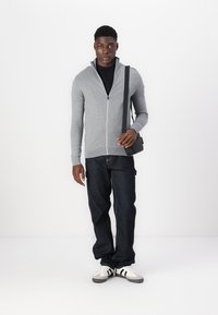 Cardigan cinza claro com fecho éclair, feito de material de malha, combinado com uma t-shirt preta e jeans escuros. Os acessórios incluem uma pequena bolsa de ombro.