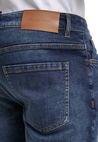 Jeans de mezclilla azul oscuro con costuras naranjas y una etiqueta de cuero marrón en la parte trasera de la cintura que dice "2V PREMIUM".