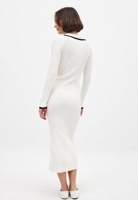 Mekko, jossa on hienot poimutukset, valkoista väriä, pitkät hihat ja musta väritys kauluksessa ja hihojen suutuksissa. Sopiva malli, joka ulottuu polven alapuolelle. Silkkinen pinta.