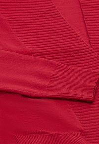 Červený pletený cardigan s kombinací hladkých a žebrovaných textur, s hlubokým V-výstřihem a dlouhými rukávy. Materiál je měkký a útulný.