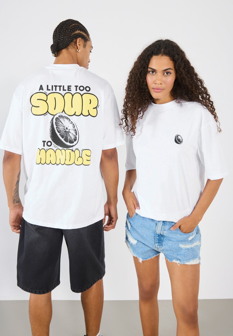 Hvid T-shirt med stor gul tekst og lime grafik på ryggen; kombineret med mørke shorts og slidte denimshorts. Sort logo på fronten.