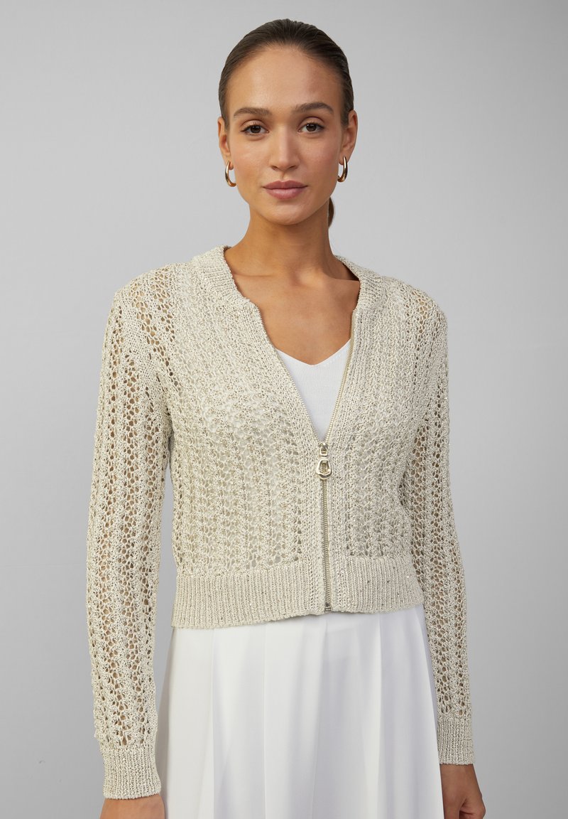 Licht beige gebreide cardigan met een rits aan de voorkant, kenmerken een gestructureerd, open geweven ontwerp en geribde manchetten en zoom.