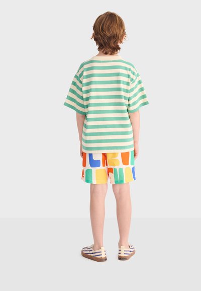 Kind steht mit dem Rücken zur Kamera, trägt ein grün-weiß gestreiftes kurzärmeliges Hemd, farbenfrohe gemusterte Shorts und gestreifte Slip-on-Schuhe.