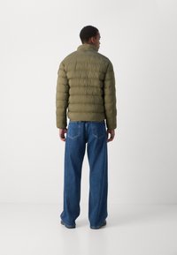 Olivengrøn pufferjakke med høj hals, kviltet struktur og elastisk kant, parret med brede blå denimjeans.