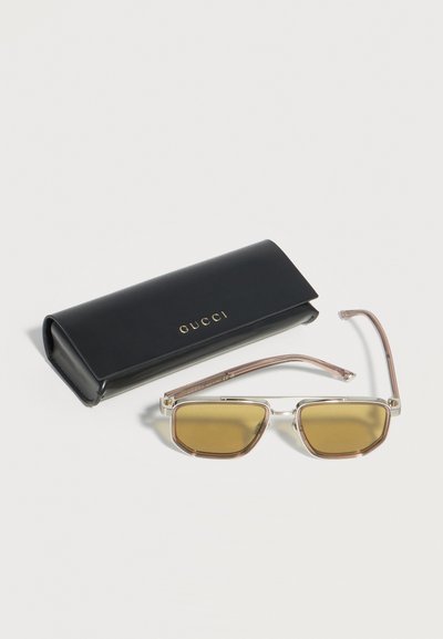Ορθογώνια γυαλιά ηλίου με κίτρινο φίλτρο και διάφανα πλαίσια δίπλα σε μαύρη θήκη Gucci με χρυσά γράμματα πάνω σε λευκή επιφάνεια.