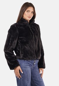 Giubbotto corto di pelliccia ecologica nera con colletto alto, chiusura con zip e texture morbida, abbinato a jeans azzurri per contrasto.