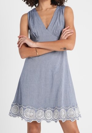 Femme portant une robe bleue sans manches avec un col en V et un ourlet brodé blanc, les bras croisés montrant un bracelet et un petit tatouage au bras.