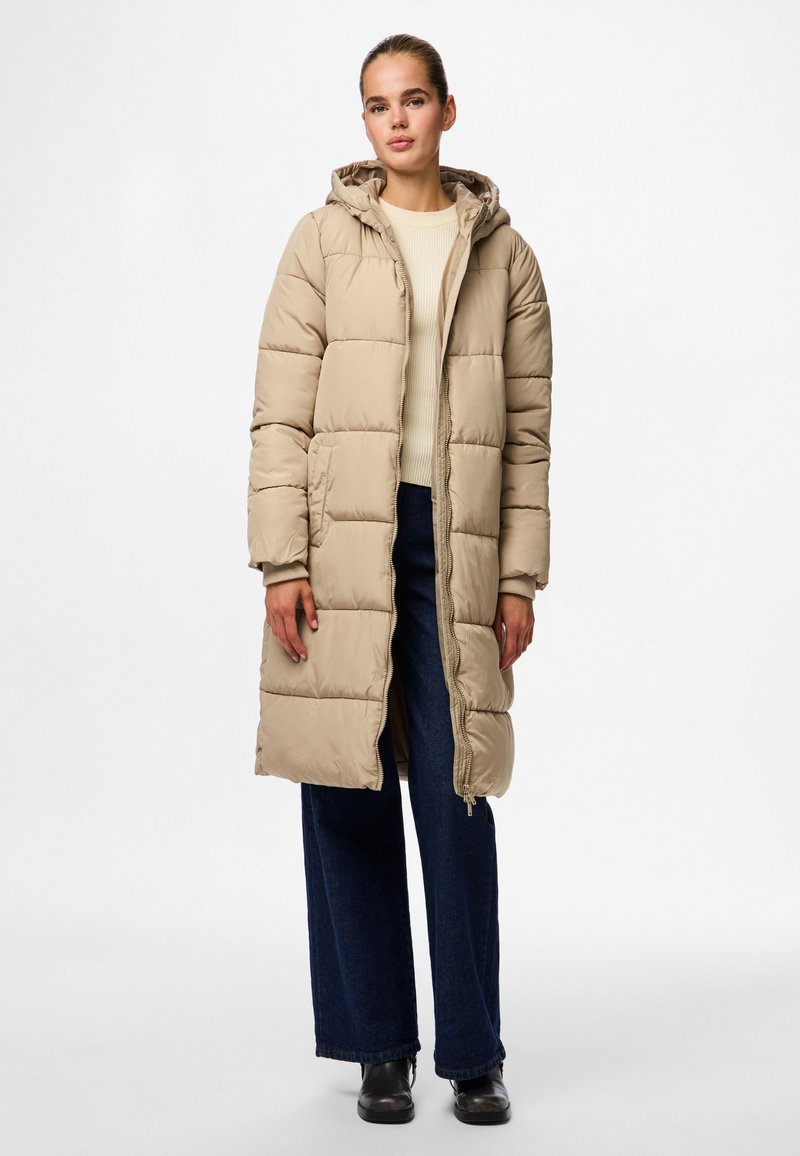 Beige lang pufferjakke med hette, quiltet design, frontglidelås og sidelommer. Kombinert med en lys genser og vide, mørke jeans.