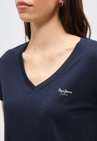 Γυναίκα που φορά μπλε σκούρο T-shirt με V λαιμόκοψη και μικρό λευκό λογότυπο "Pepe Jeans London" στο αριστερό στήθος, ορατή από το πηγούνι έως το μέσο του κορμού.