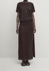 Top in lino marrone con maniche arrotolate, abbinato a una gonna avvolgente marrone con uno spacco laterale. Indossato con sandali neri con cinturini.