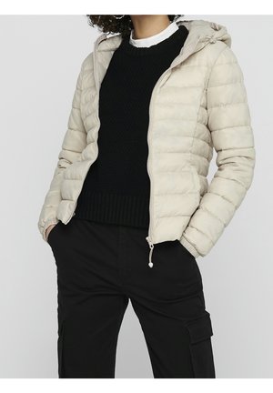 Person trägt eine beige gesteppte Kapuzenjacke über einem schwarzen Pullover und schwarzen Hosen, die Hände in den Taschen, vor einem schlichten Hintergrund.