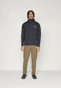 Veste noire zippée avec capuche, en tissu léger. Pantalons vert olive, coupe ajustée. Les deux présentent des accents de logo. Des chaussures de sport noires complètent l'ensemble.