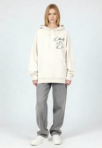 Felpa con cappuccio color crema con una tasca frontale, decorata con un disegno a contorno nero di un uccello e la parola "Liberté"; abbinata a jeans grigi a vestibilità ampia e scarpe da ginnastica bianche.