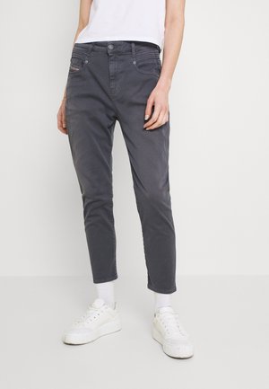 Persoon draagt donkergrijze slim-fit broek, witte sneakersokken, witte sneakers en een wit T-shirt met korte mouwen, staand tegen een effen achtergrond.