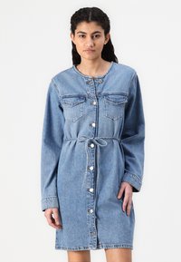 Denim obleka v svetlo modri barvi, z dolgimi rokavi, gumbasto zaponko spredaj, dvema žepoma na prsih, pasom na vrvico in ravnim krojem.