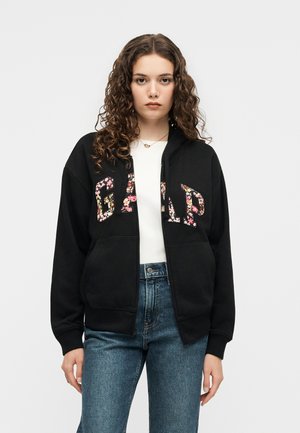 Jeune femme aux cheveux bouclés portant un sweat à capuche noir zippé avec les lettres « GAP » fleuries sur un haut blanc et un jean bleu, debout devant un fond uni.