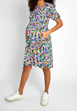 Kobieta w ciąży stoi z rękami w kieszeniach, ubrana w kolorową kwiecistą sukienkę do kolan i białe tenisówki na jednolitym tle.