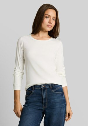 Junge Frau mit langen braunen Haaren, die ein weißes langärmeliges Shirt und blaue Jeans trägt, steht vor einem schlichten hellgrauen Hintergrund.