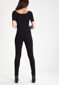 Kvinde står med ryggen til, iført en sort kortærmet top, sorte tætsiddende jeans og sorte ankelstøvler med blokhæl.