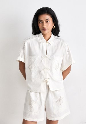 White cotton shirt and shorts set featuring lace accents and embroidered patches, with a tie closure and short sleeves.