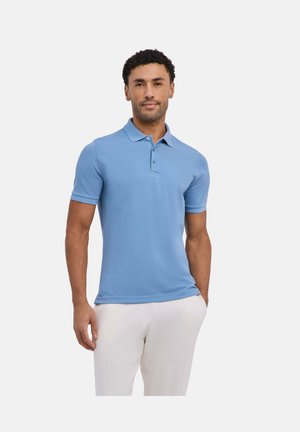 Mann mit kurzen lockigen Haaren, der ein hellblaues kurzärmeliges Poloshirt und weiße Hosen trägt, vor einem schlichten Hintergrund stehend.