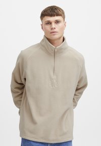 Beige fleece pullover with a half-zip collar, featuring a textured circular logo at the center. Soft fabric and relaxed fit.