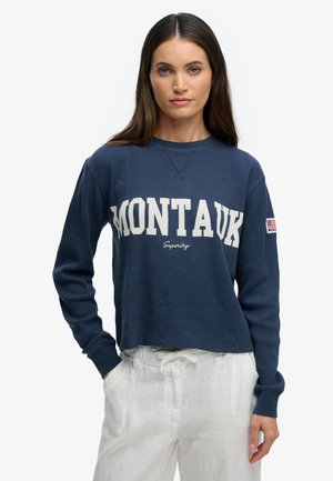 Kvinde iført en mørkeblå Montauk sweatshirt med hvide bogstaver og hvide bukser, stående med den ene hånd i lommen mod en hvid baggrund.