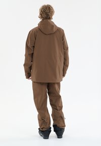 Person står med ryggen til iført en brun hættejakke og matchende bukser med sorte sneboots på hvid baggrund.