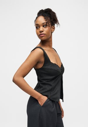 Młoda kobieta z kręconymi włosami związanymi w wysoki kucyk, ubrana w czarny bezrękawnik w cienkie paski i ciemne spodnie, pozująca z jedną ręką w kieszeni.