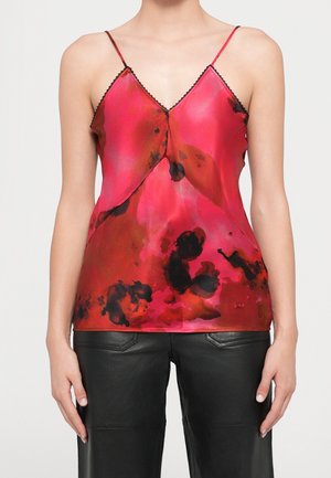 Kvinde iført en rød og sort asymmetrisk ærmeløs top med tynde stropper, sat sammen med sorte læderbukser mod en neutral baggrund.