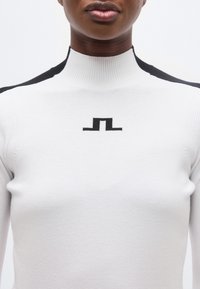 Κοντινό πλάνο ενός ατόμου που φοράει λευκό πουλόβερ με ψηλό γιακά, με μαύρες λεπτομέρειες στους ώμους και μαύρο γεωμετρικό λογότυπο στο στήθος.